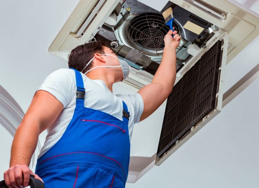T&eacute;cnico con mono azul reparando un aire acondicionado de techo con un destornillador.